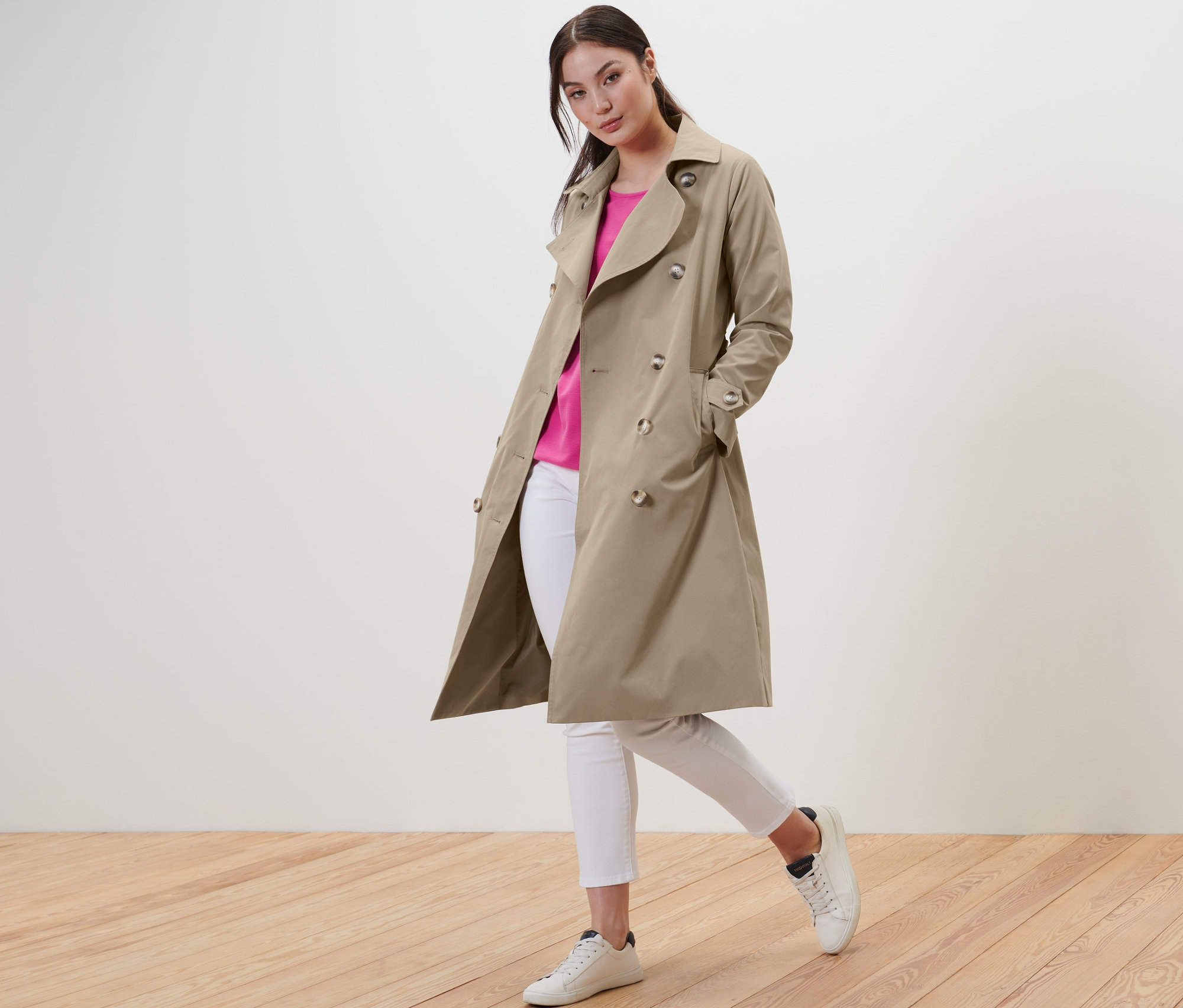 Frau posiert in beigem Trenchcoat, weißen Jeans und Turnschuhen.