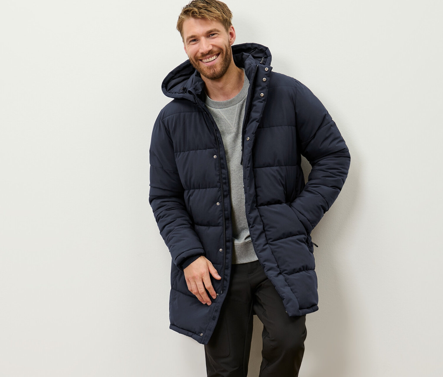 Ein lächelnder Mann lehnt an einer Wand und trägt einen Stepp-Parka mit Kapuze.