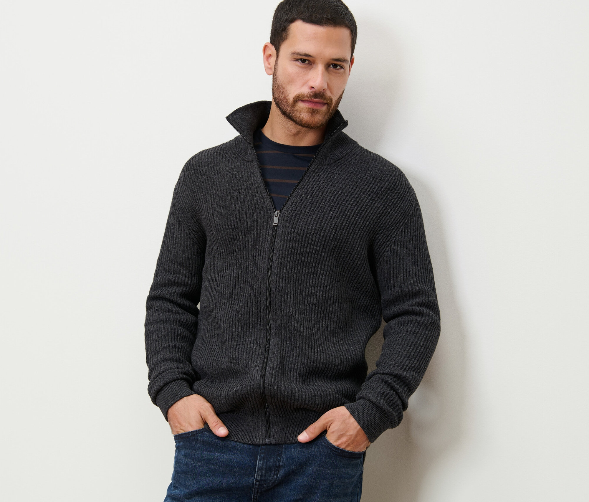 Mann steht mit Händen in den Hosentaschen und trägt eine melierte anthrazitfarbene Strickjacke.