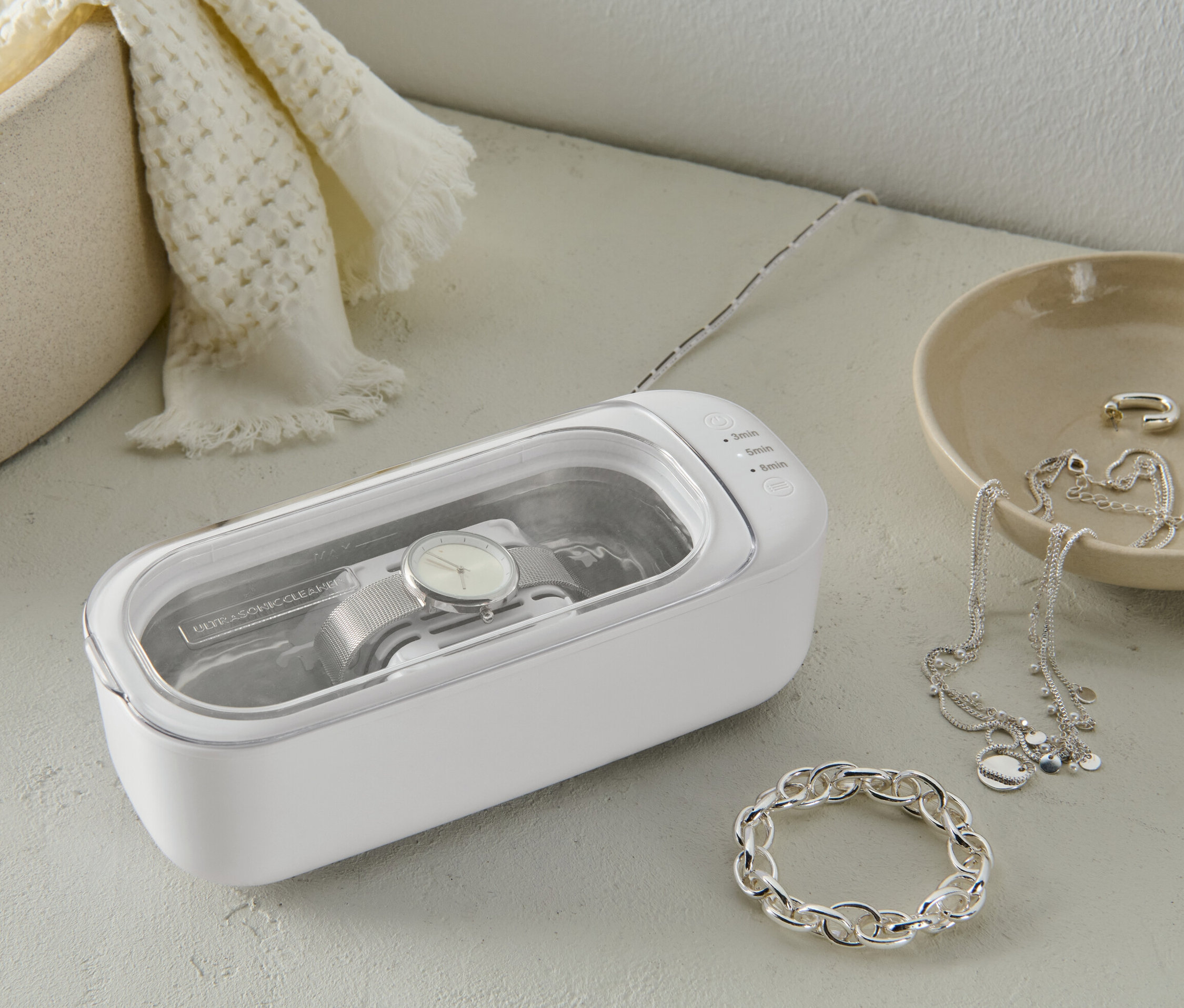 Eine Uhr wird in einem weißen Ultraschallreiniger gereinigt. Daneben liegt Schmuck.
