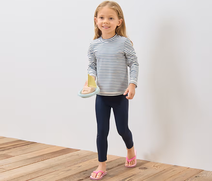 Mädchen hält Spielzeugboot, trägt Kinder-Shirt mit UV-Schutzfaktor 80, Leggings und Zehentrenner.
