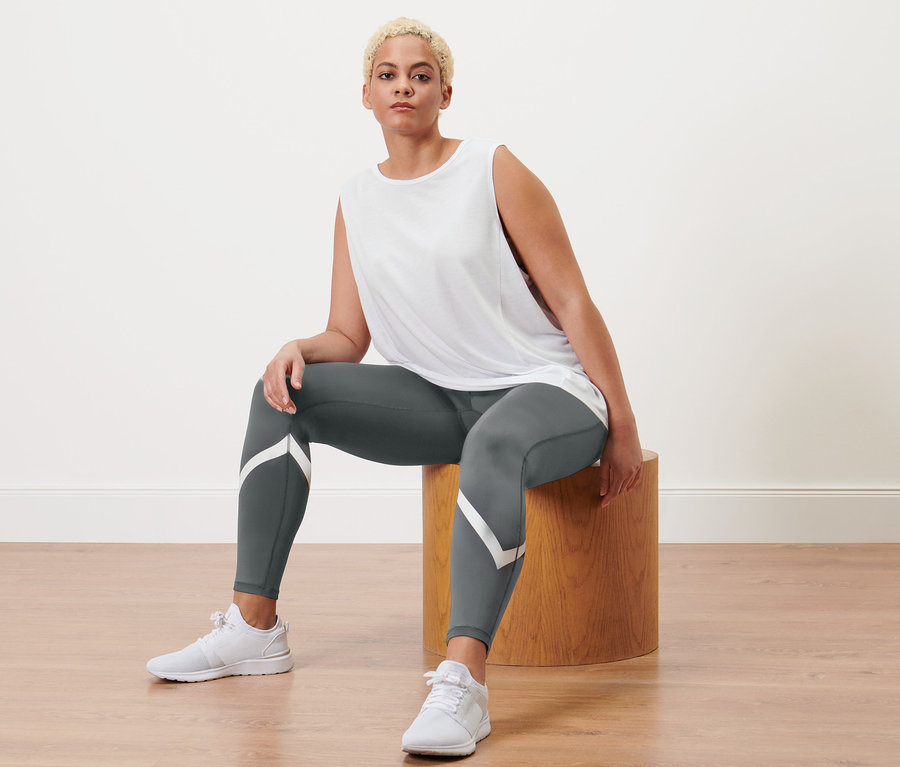 Frau sitzt auf einem Holzhocker, trägt graue Sporttight und 2 Sport-Tops, grau und weiß.
