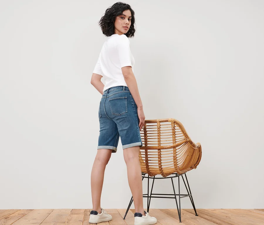 Frau posiert in Bermuda Jeans – Fit »Lea« und weißem T-Shirt neben einem Korbstuhl.