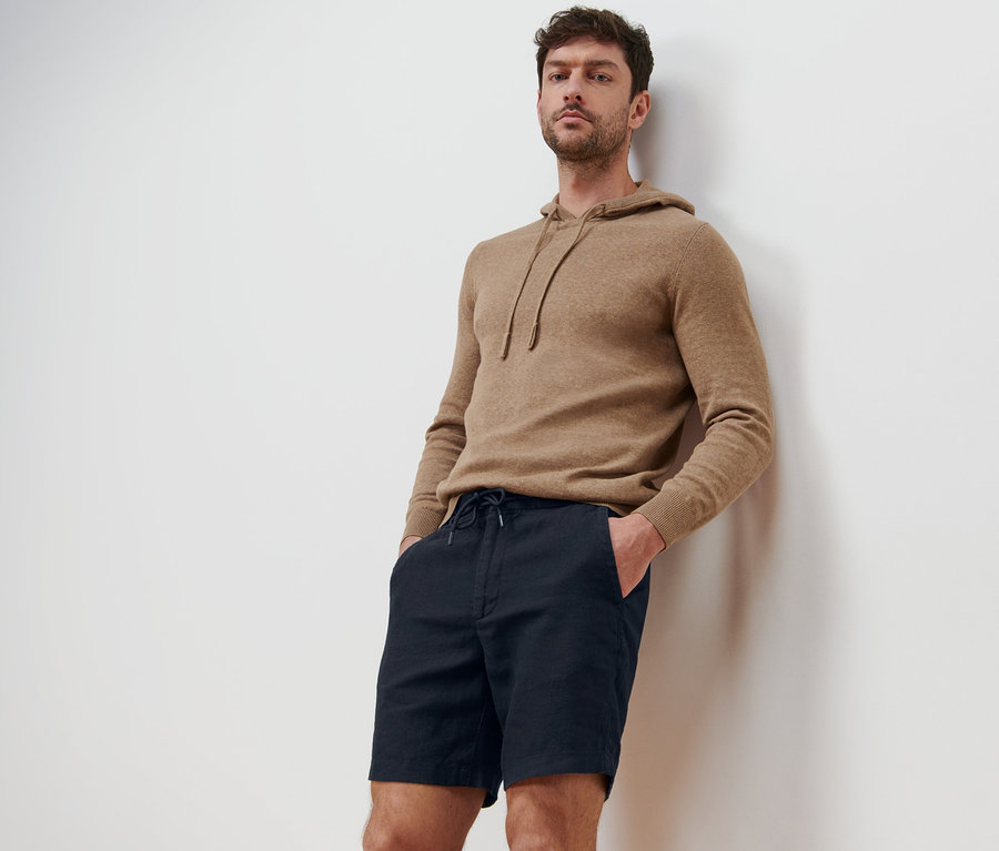 Mann lehnt an einer Wand und trägt Leinen-Shorts und einen Kapuzenpullover.