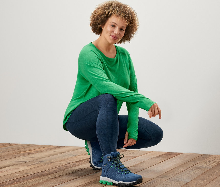Frau in grünem Langarm-Sportshirt und Sporttights posiert auf einem Holzboden.