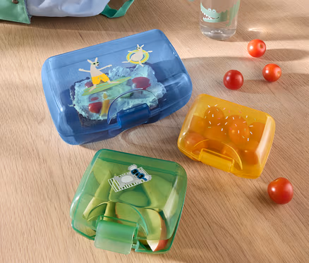 Drei Brotdosen liegen auf einem Tisch, zusammen mit einer Wasserflasche und einem Rucksack.