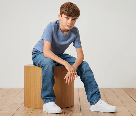 Junge sitzt auf einem Holzhocker und trägt blaue Kinder-Jeans – Fit »Jonas« und ein blaues T-Shirt.