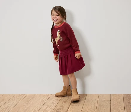 Ein lächelndes Mädchen mit Zöpfen trägt einen bordeauxroten Pullover mit einem Pferd, einen Rock und braune Stiefel.