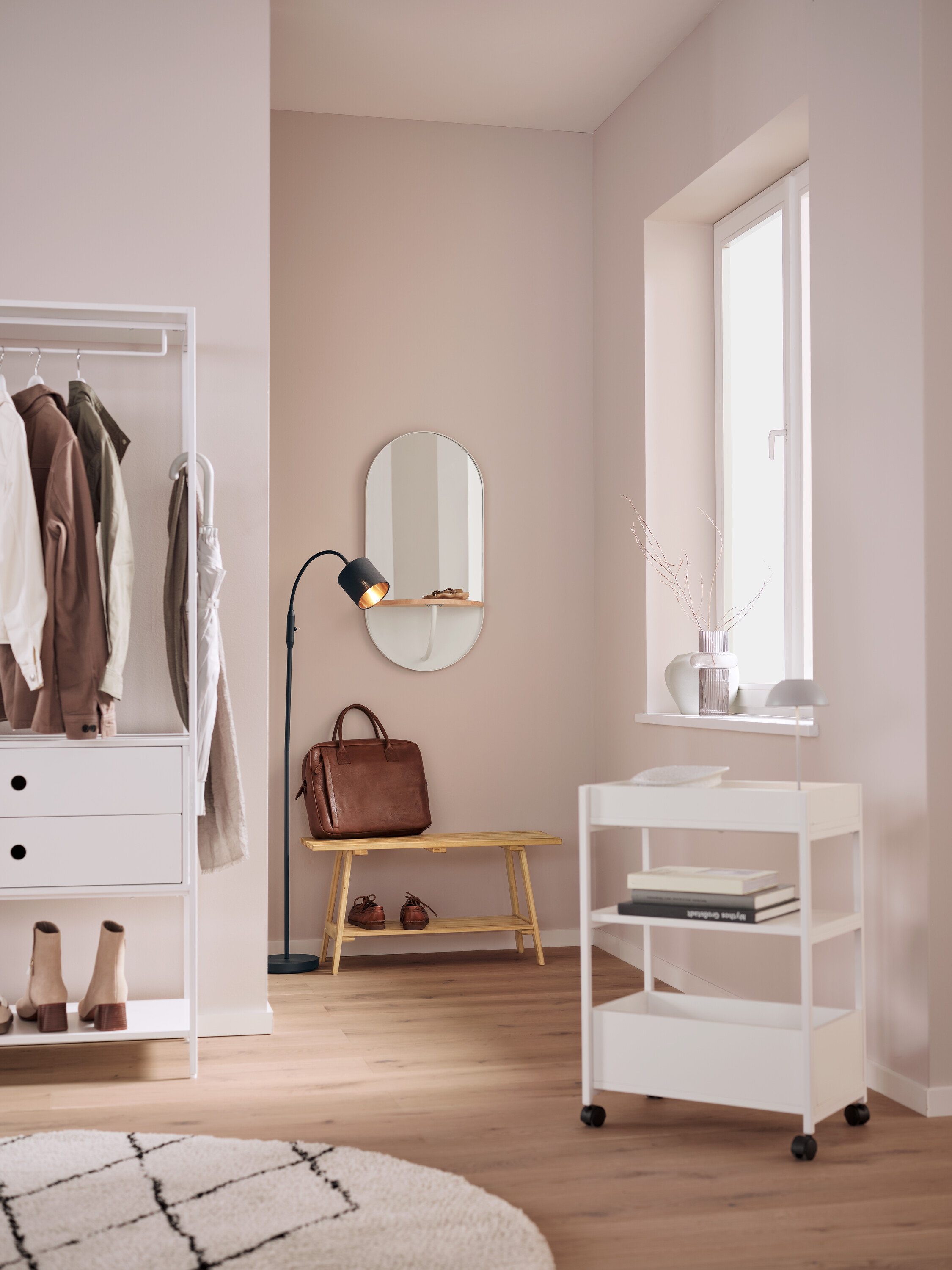 In einem Interieur befinden sich ein weißes Regal mit Kleidung, ein ovaler Spiegel, eine braune Ledertasche auf einer Bank, eine schwarze Lampe und ein weißer Rollwagen.