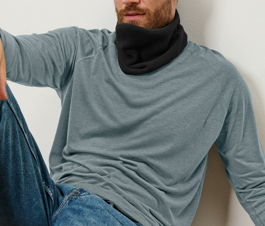 Ein Mann sitzt angelehnt an einer Wand und trägt einen schwarzen Nackenwärmer, blaue Jeans und ein blau-graues Langarmshirt.