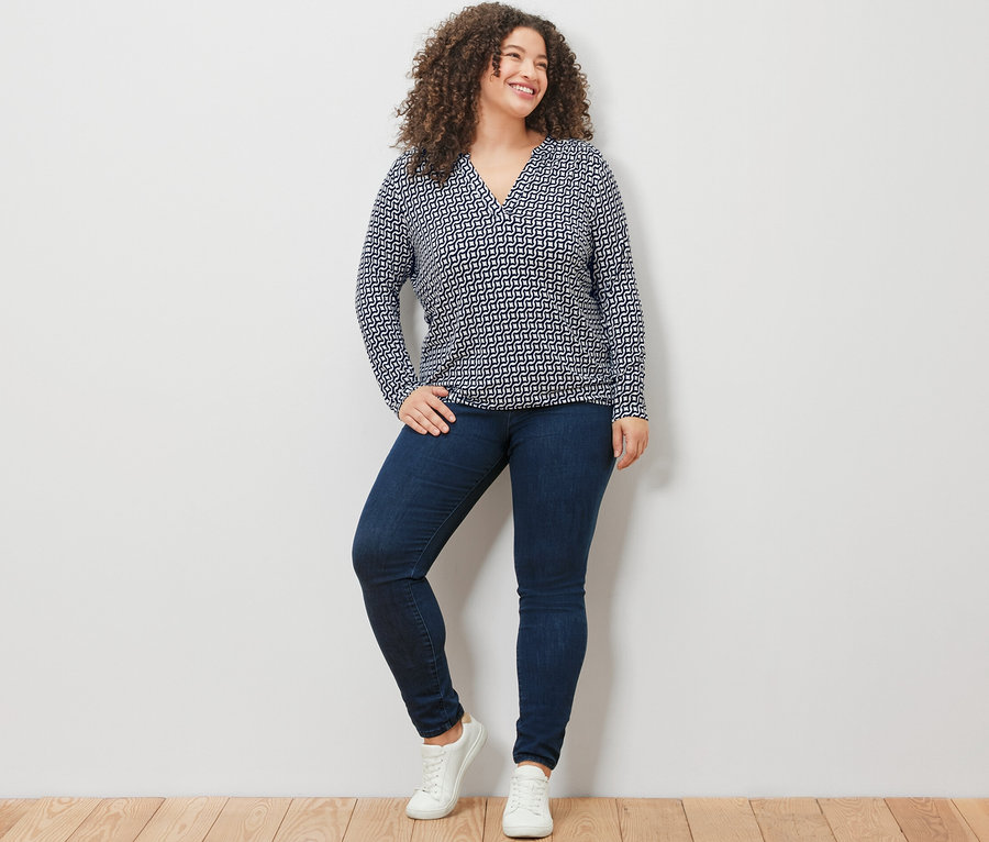 Frau steht vor einer weißen Wand und trägt ein bedrucktes Blusenshirt in Navy, Jeans und weiße Turnschuhe.