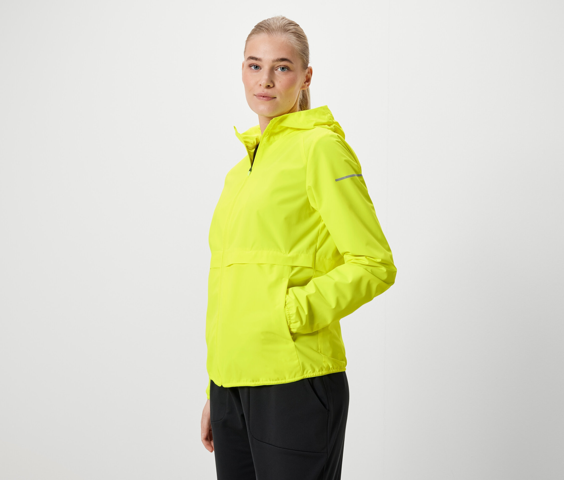 Blonde Frau in neongelber Kapuzenjacke und schwarzen Hosen posiert seitlich.