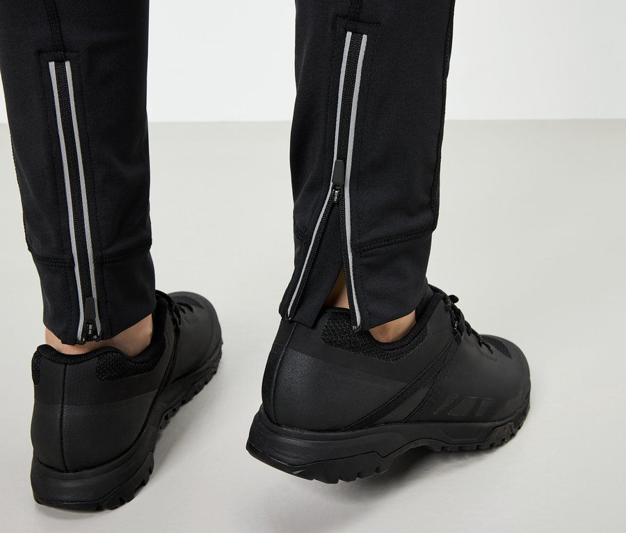 Beine in schwarzen Sportschuhen und schwarzen Hosen mit silbernen Reißverschlüssen auf grauem Hintergrund.