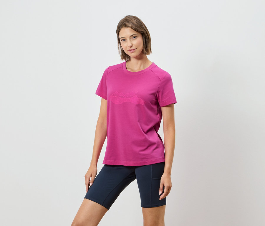 Frau trägt ein pinkes T-Shirt mit einer Bergkette und dunkelblaue Shorts vor einem weißen Hintergrund.