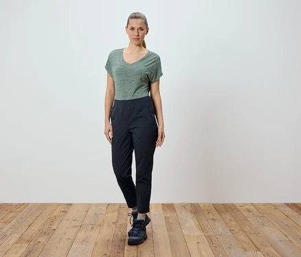 Eine Frau steht vor einer weißen Wand auf einem Holzboden. Sie trägt ein grünes T-Shirt und eine schwarze Hose.