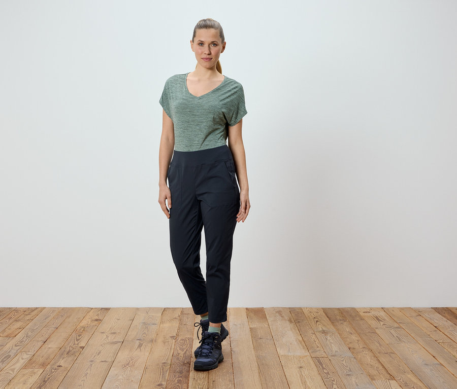 Eine Frau steht vor einer weißen Wand auf einem Holzboden. Sie trägt ein grünes T-Shirt und eine schwarze Hose.
