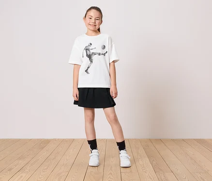 Ein junges Mädchen trägt ein weißes T-Shirt mit einem Fußballspieler-Motiv, einen schwarzen Rock, Socken und weiße Schuhe.