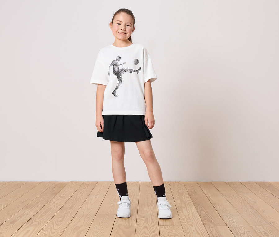 Ein junges Mädchen trägt ein weißes T-Shirt mit einem Fußballspieler-Motiv, einen schwarzen Rock, Socken und weiße Schuhe.