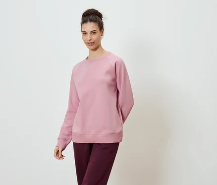 Frau trägt ein rosafarbenes Sweatshirt und eine burgunderfarbene Hose vor einem weißen Hintergrund.