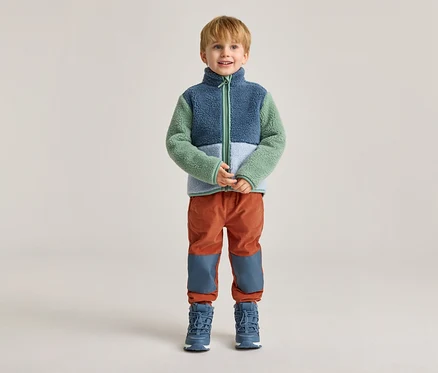 Ein Junge mit blonden Haaren steht vor einem hellen Hintergrund in einer blau-grün-weißen Fleecejacke, orangefarbenen Hosen und blauen Stiefeln.