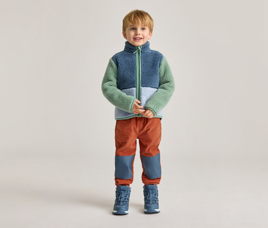 Ein Junge mit blonden Haaren steht vor einem hellen Hintergrund in einer blau-grün-weißen Fleecejacke, orangefarbenen Hosen und blauen Stiefeln.