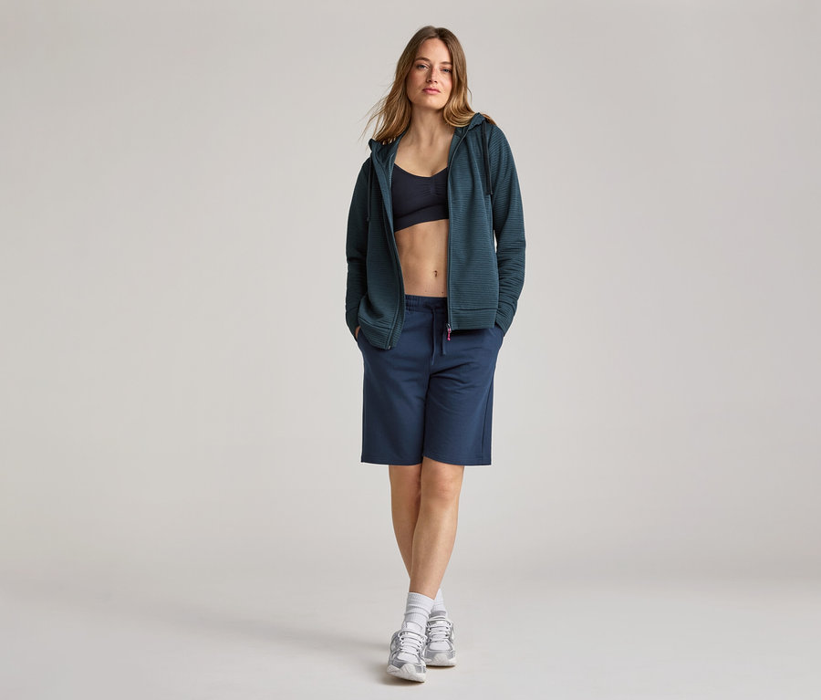 Frau mit marineblauer Kapuzenjacke, Shorts und Sportschuhen. Sie trägt eine Kapuzenjacke, navy, offen über einem Sport-BH.