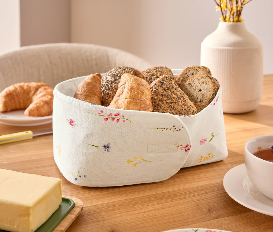 Auf einem Holztisch steht ein Brotkorb mit verschiedenen Brotsorten, darunter Croissants und Brötchen, neben einer Tasse Tee, einem Stück Butter und einem Teller mit Blumenmuster.