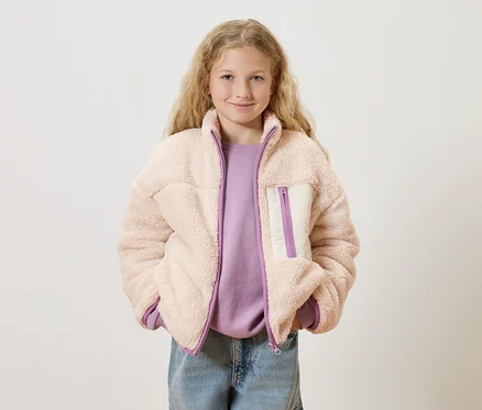 Mädchen mit blonden, lockigen Haaren trägt eine rosa Teddyfelljacke über einem lila Pullover und blauen Jeans.