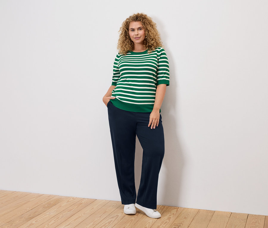 Frau mit lockigem Haar trägt ein grün-weiß gestreiftes Oberteil, dunkelblaue Hosen und weiße Schuhe.