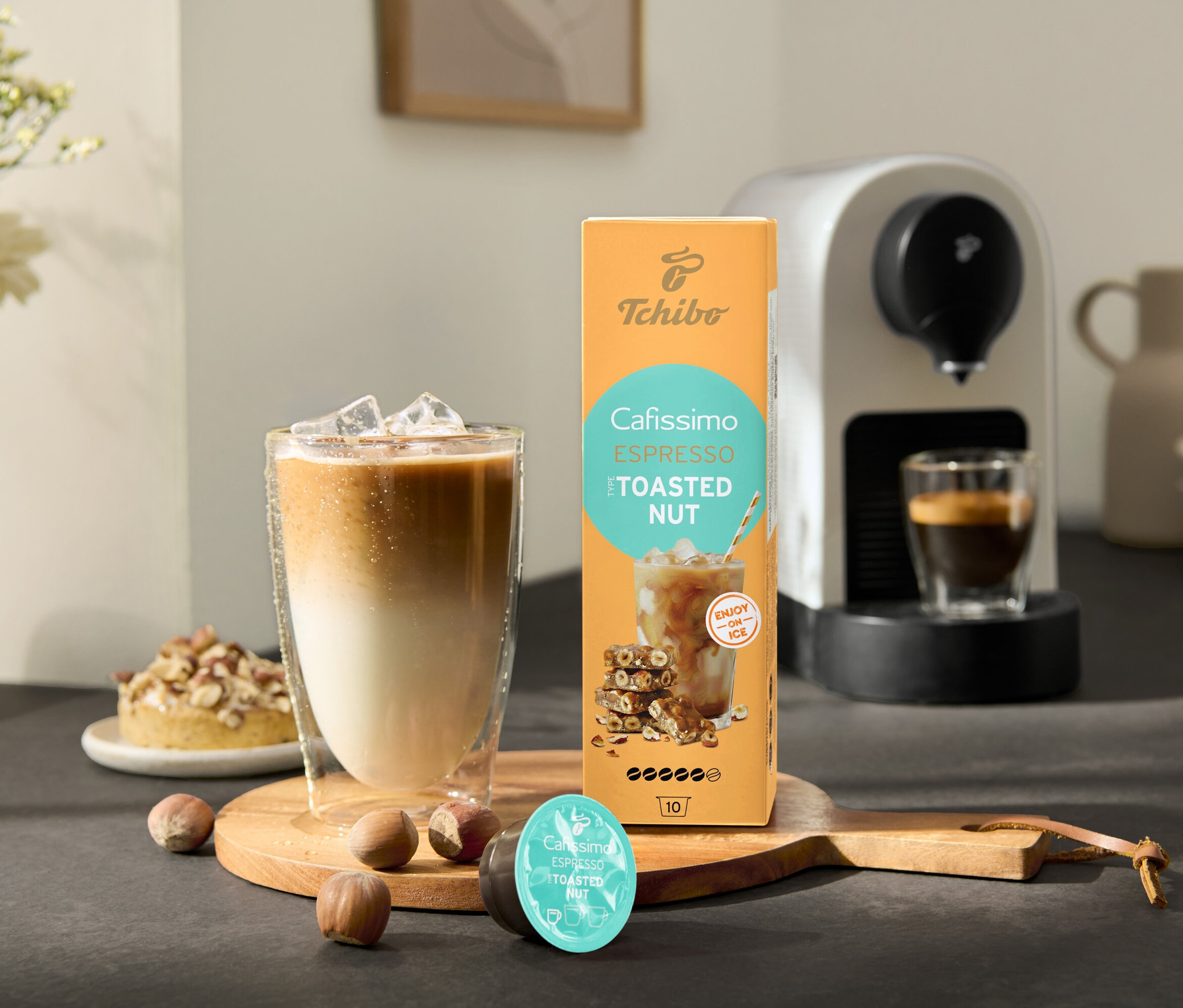 Ein Glas Eiskaffee steht neben einer Packung Cafissimo Flavoured Espresso – Toasted Nut und einer Cafissimo pure+, white Kaffeemaschine.