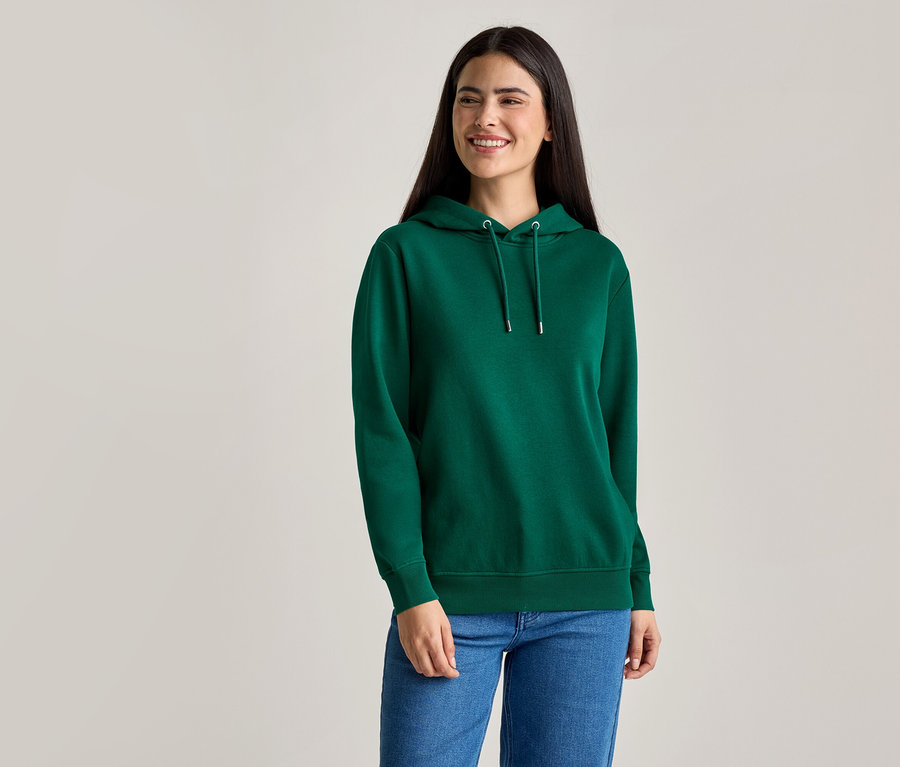 Frau mit langen, dunklen Haaren trägt einen grünen Kapuzenpullover und blaue Jeans.