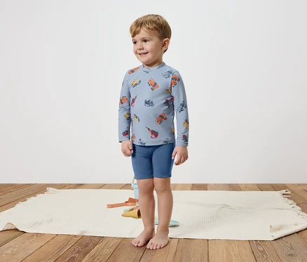 Ein kleiner Junge steht barfuß auf einem Teppich und trägt ein blaues, langärmliges Oberteil mit bunten Fahrzeugen und blaue Shorts.