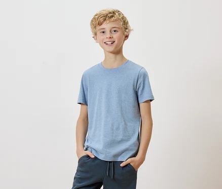 Ein blonder Junge mit einem hellblauen T-Shirt und dunklen Hosen steht vor einem weißen Hintergrund.