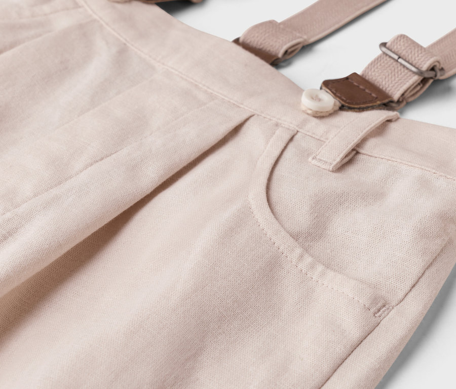 Detailaufnahme einer hellbeigen Hose mit Hosenträgern, Knopf und Tasche.
