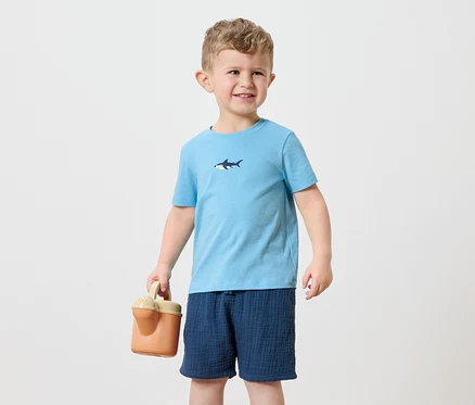 Ein kleiner Junge mit blonden Haaren trägt ein hellblaues T-Shirt mit einem Hai-Aufdruck und marineblaue Shorts. Er hält eine orangefarbene Gießkanne.