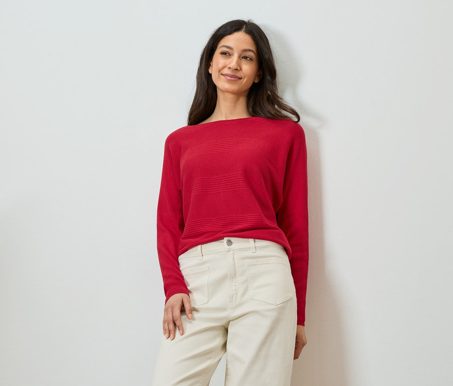 Frau lehnt an einer Wand, trägt einen roten Pullover und eine beigefarbene Hose.