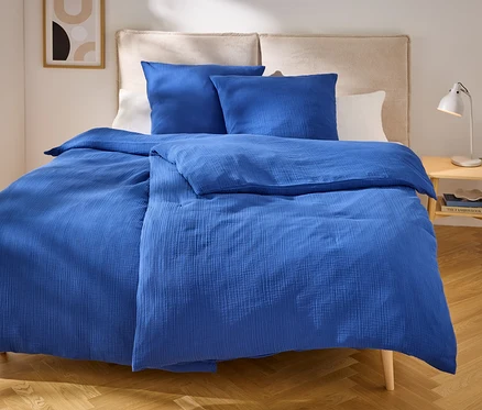 Blaues Bettzeug mit zwei Kissen auf einem Bett mit beigem Kopfteil. Auf dem Nachttisch steht eine Lampe.