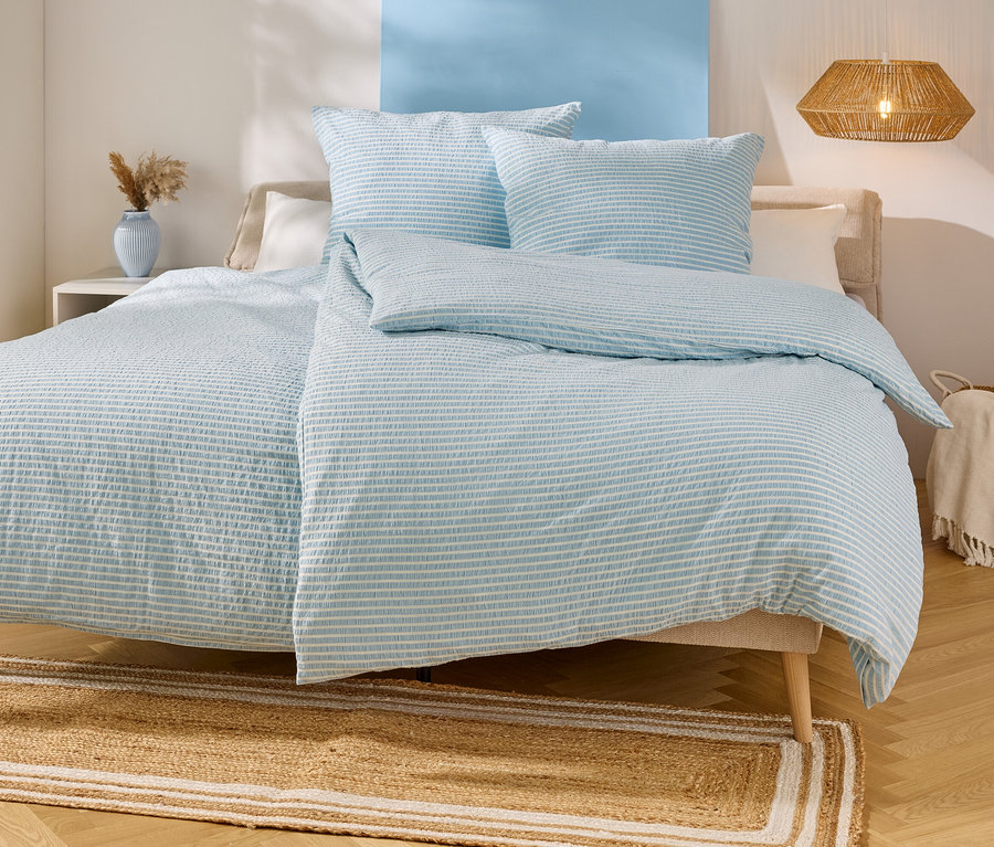 Bett mit hellblau-weiß gestreifter Bettwäsche, zwei Kissen und einer Decke. Ein beiger Teppich liegt unter dem Bett.