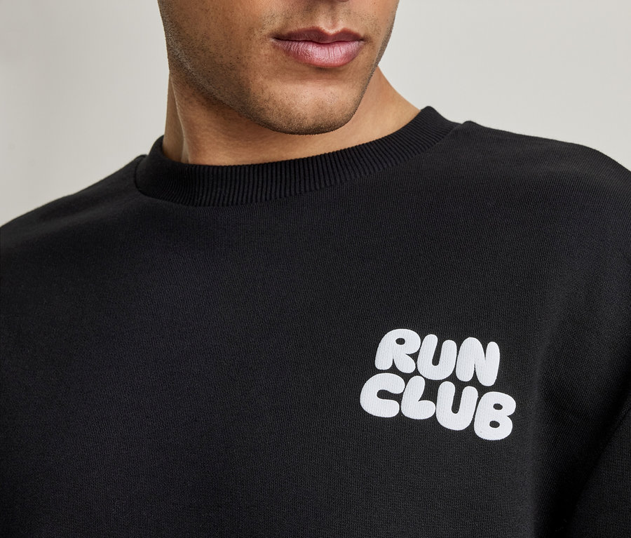 Nahaufnahme eines Mannes, der ein schwarzes Sweatshirt mit dem weißen Schriftzug 'RUN CLUB' trägt.