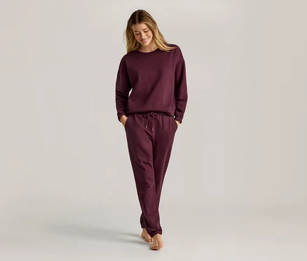 Frau mit langen blonden Haaren, die ein weinrotes, langärmeliges Sweatshirt und eine Jogginghose trägt und barfuß geht.