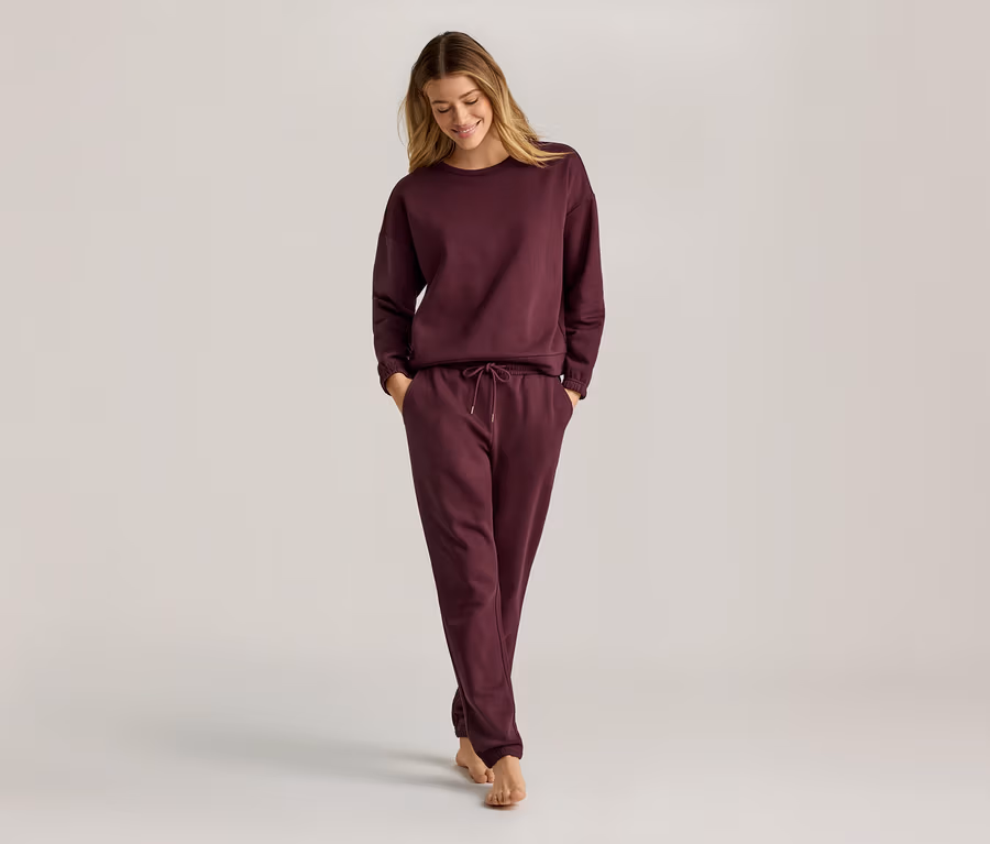 Frau mit langen blonden Haaren, die ein weinrotes, langärmeliges Sweatshirt und eine Jogginghose trägt und barfuß geht.