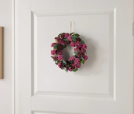 Kleiner Blumenkranz mit lila Blüten und grünen Blättern hängt an einer weißen Tür.