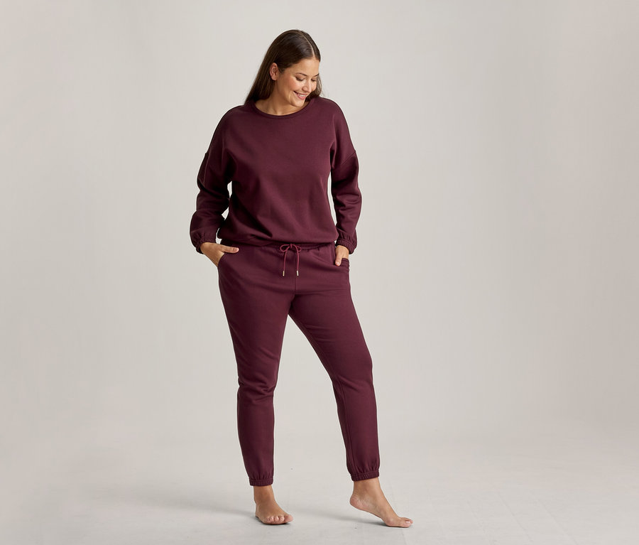 Eine lächelnde Frau steht und blickt nach unten, trägt ein bordeauxrotes Loungewear-Set aus Sweatshirt und Jogginghose. Ihre Hände stecken in den Hosentaschen, und sie ist barfuß.