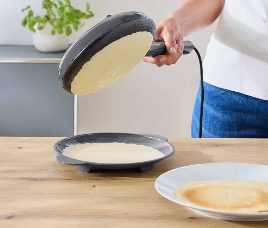 Eine Person neigt eine dunkelgraue elektrische Bratpfanne über einen Teller mit restlichem Teig. Ein fertiger Crêpe liegt auf einem weißen Teller auf einer Holzablage.