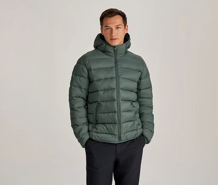 Mann in einer grünen Kapuzen-Steppjacke und dunklen Hosen, der mit den Händen in den Taschen steht.