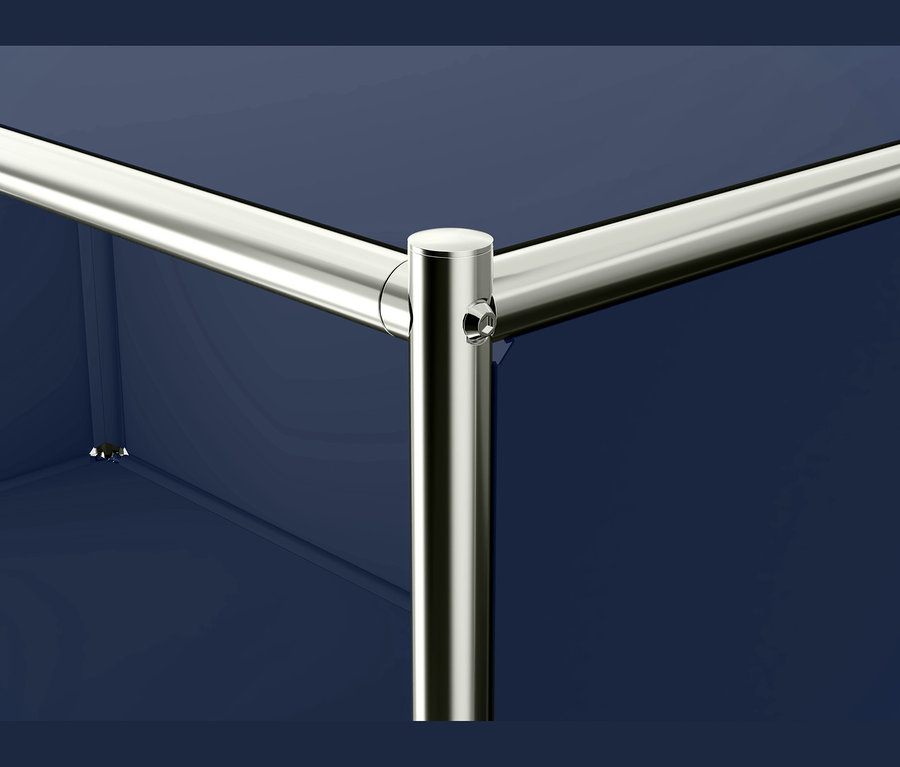 Nahaufnahme einer modularen Möbelverbindung, die drei verchromte Metallrohre zeigt, die an einem zentralen Verbinder mit sichtbarer Schraube vor einer dunkelblauen Platte zusammenlaufen.