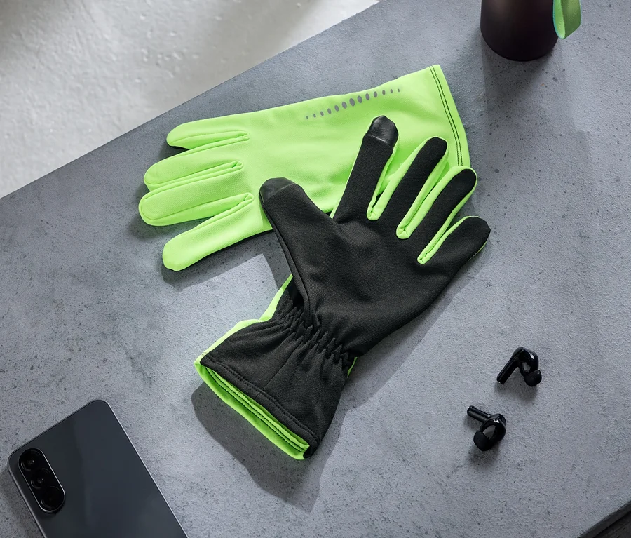 Ein Paar neongrüne und schwarze Handschuhe mit Touchscreen-Fingerspitzen liegt auf einer grauen Oberfläche neben einem dunklen Smartphone und zwei schwarzen kabellosen Ohrhörern. Eine dunkle Flasche ist teilweise sichtbar.