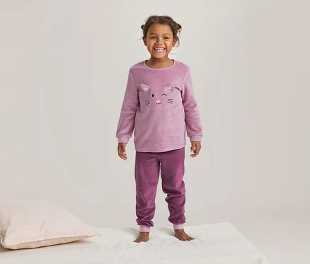 Lächelndes kleines Mädchen in einem rosa Pyjama mit Katzenmotiv und gestreiften Hosen steht auf einer weißen Decke neben einem rosa Kissen.