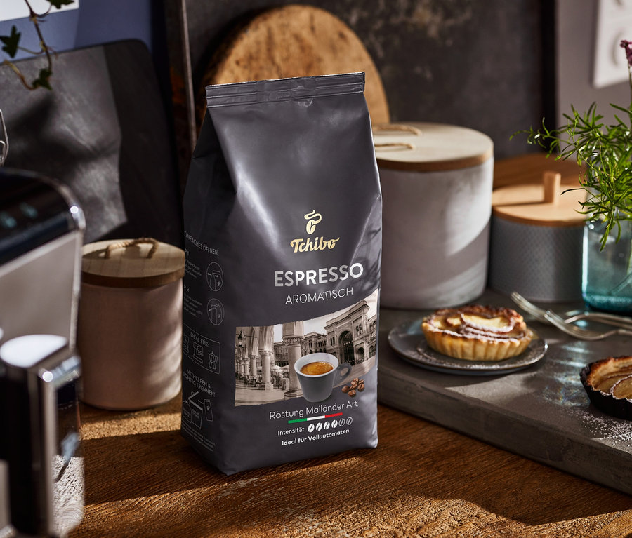 Eine Packung Espresso Aromatisch steht auf einem Holztisch neben einer Kaffeemaschine und Tellern mit Kuchen.