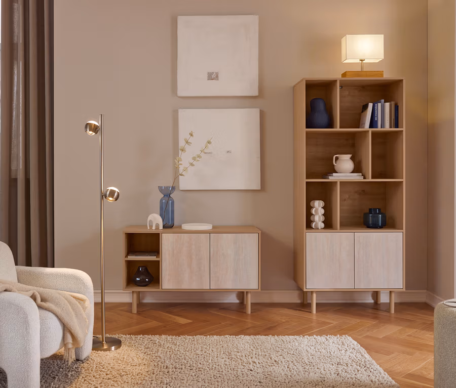Gemütliches Wohnzimmer mit hellen Holzmöbeln. Ein weißer Sessel mit beiger Decke steht neben einer Messing-Stehlampe. Zwei abstrakte Bilder hängen über einem niedrigen Schrank mit blauer Vase. Ein hohes Bücherregal mit Deko und Büchern wird von einer Tischlampe gekrönt. Ein zotteliger Teppich liegt auf einem Fischgrätparkett.
